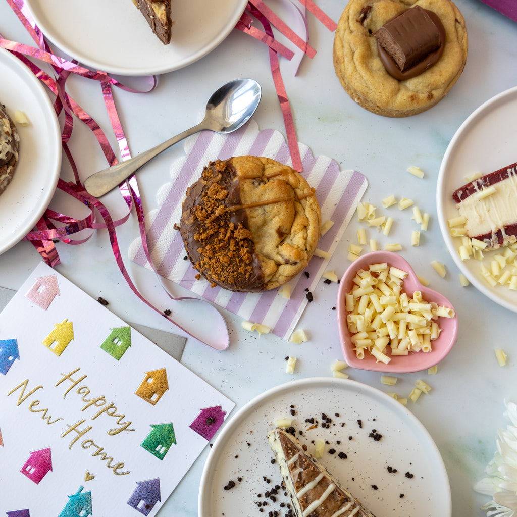 A styled flat-lay of assorted baked goods on a pale blue surface, including a half-chocolate half-brown crumb-topped pastry, a cookie with a chocolate chunk, a slice of cookie pie with white chocolate drizzle, a heart-shaped bowl of white chocolate pieces, and a 'Happy New Home' card.