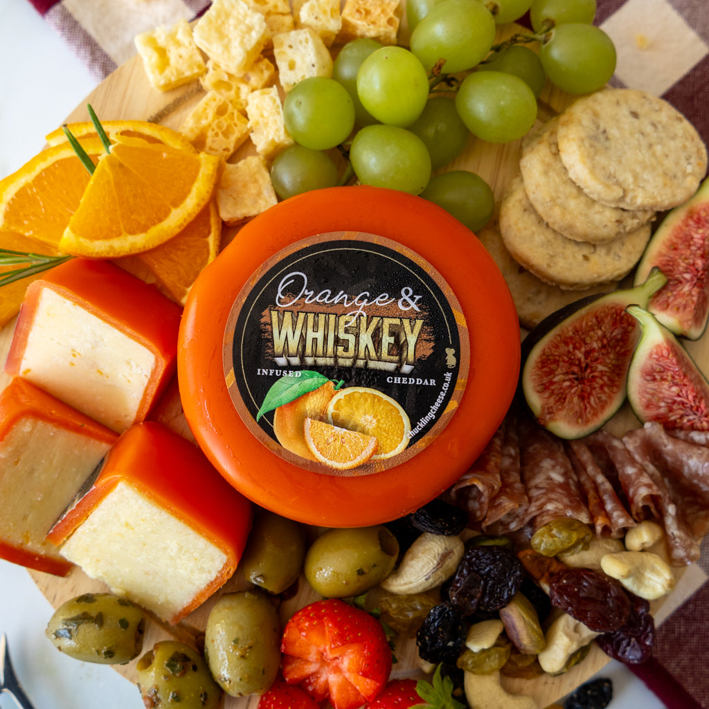 A round wheel of orange cheddar cheese labeled 'Orange's Whiskey' sits on a charcuterie board surrounded by sliced orange, green grapes, crackers, figs, olives, nuts, dried fruit, and cured meat.
