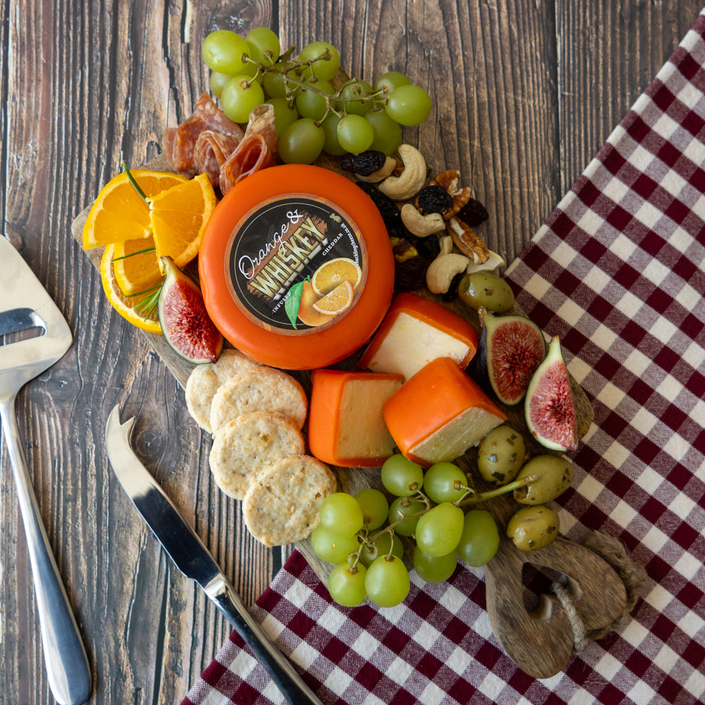 A styled charcuterie board featuring a round orange cheese labeled 'Orange & Whiskey', wedges and slices of orange cheese, crackers, green grapes, figs, cashews, dried fruit, and orange slices on a rustic wooden table with a red-and-white checkered cloth.