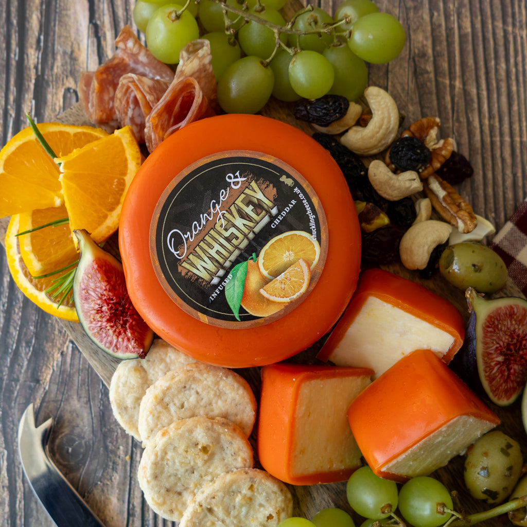 An assortment of cheeses, crackers, grapes, figs, prosciutto, and citrus arranged on a rustic wooden board, with a round orange cheese labeled 'Orange & Whiskey' prominently centered.