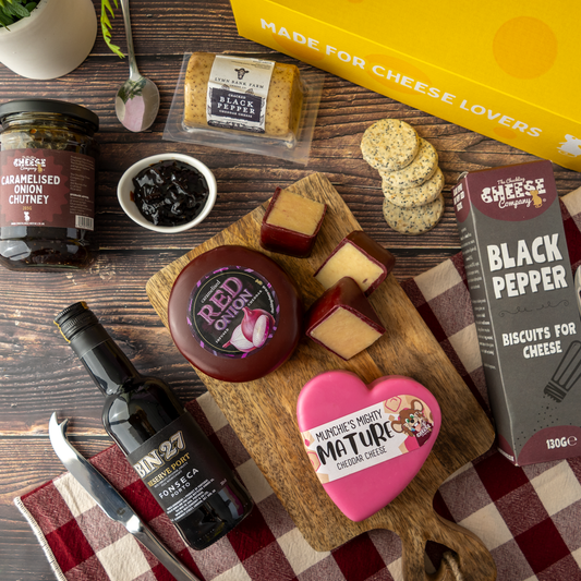A cheese and charcuterie gift box flat lay on a dark wood background, featuring a yellow 'Made for Cheese Lovers' box, a jar of caramelised onion chutney, a bottle of Bin-27, a package of black pepper biscuits, a wheel of red cheese, sliced cheeses, crackers, and a pink heart-shaped cheese tin.