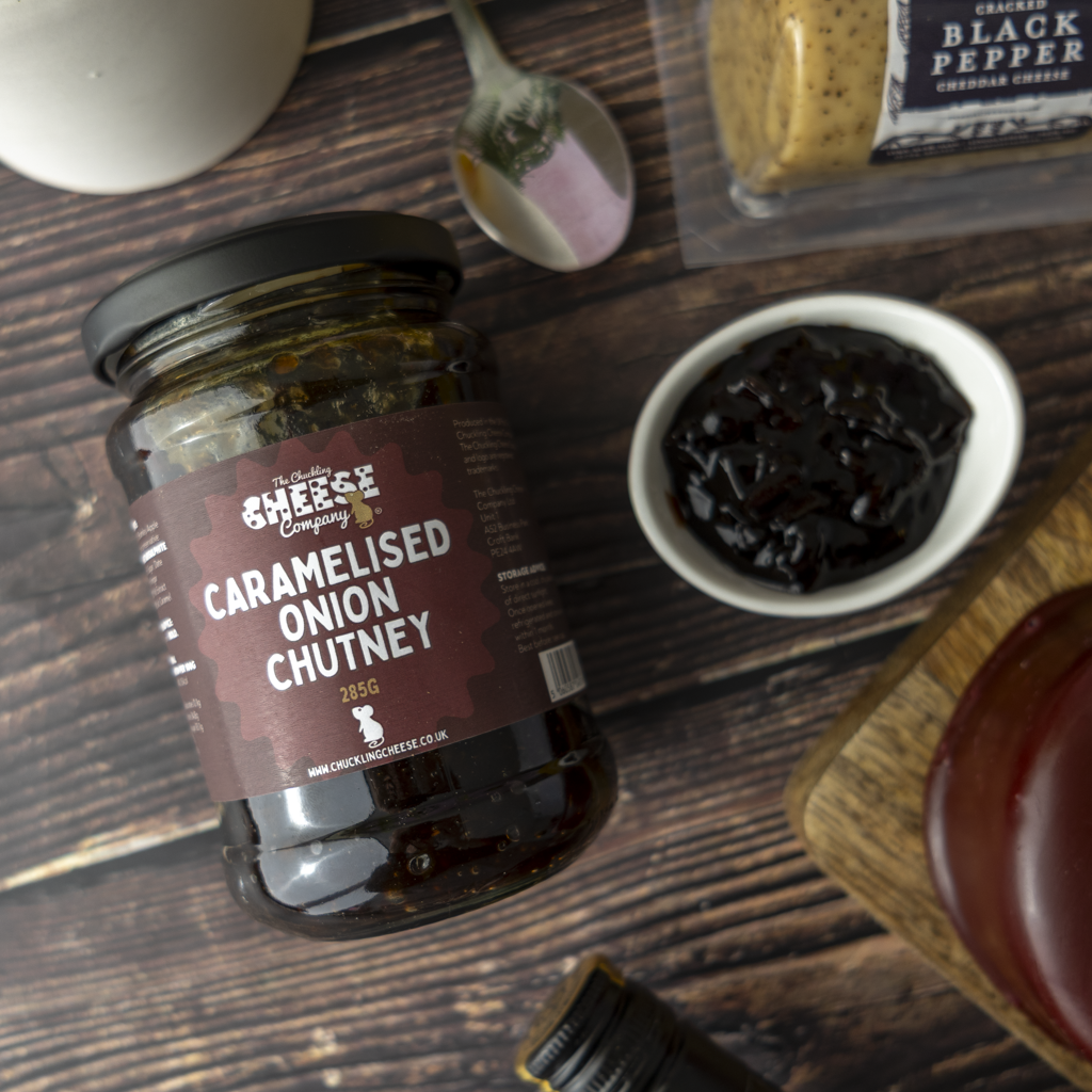 A jar of The Chuckling Cheese Company Caramelised Onion Chutney on a dark wooden tabletop, surrounded by a small bowl of dark chutney, a spoon, and other kitchen items.