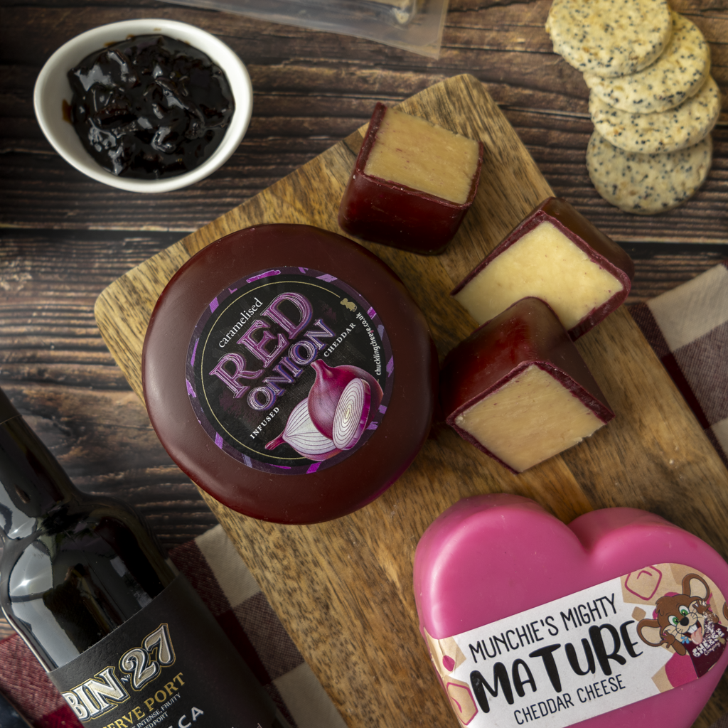 A styled food flat-lay featuring a round wheel of Caramelised Red Onion cheese, several wedges of cheddar cheese with red onion skins, a pink heart-shaped Munchie's Mighty Mature cheddar cheese box, a small bowl of dark jam, and a bottle of Bin 27 wine on a wooden board.