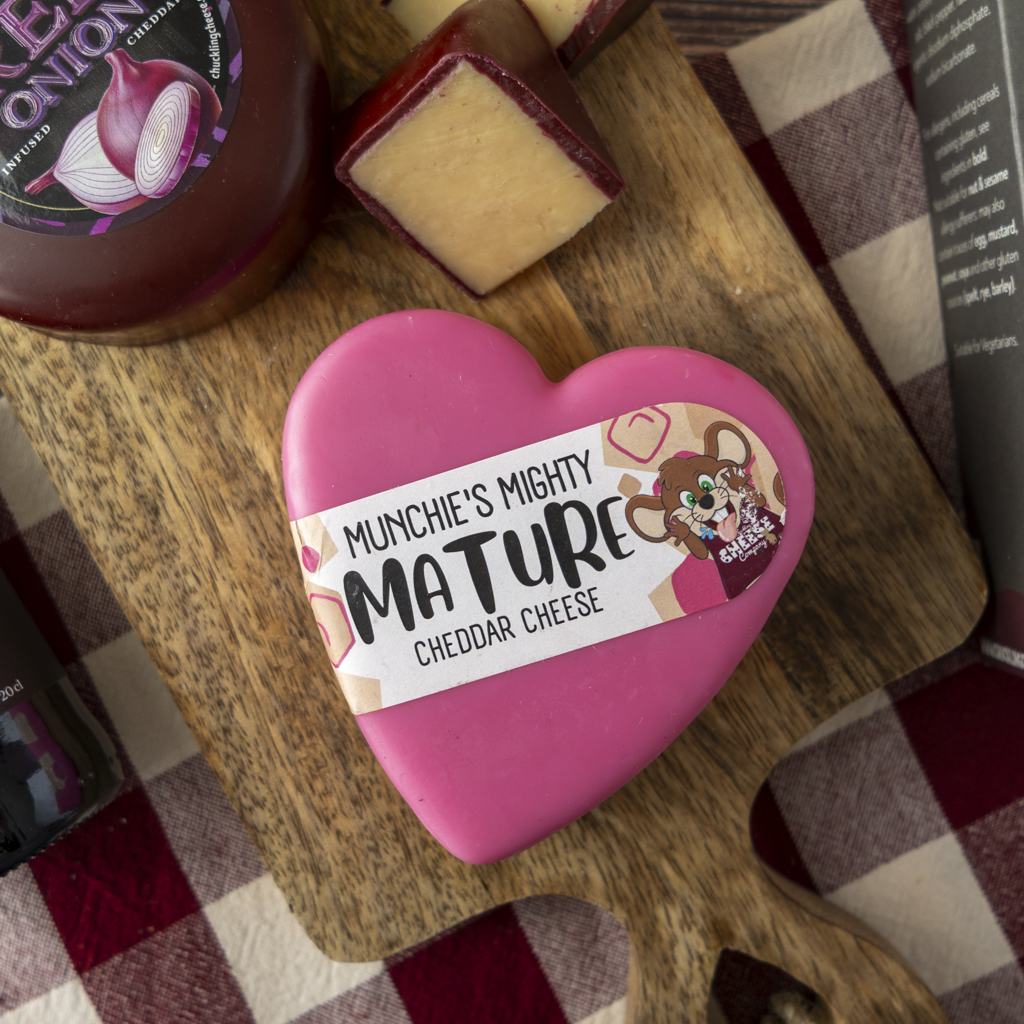 A heart-shaped pink cheese block labeled 'Munchie's Mighty Mature Cheddar Cheese' sits on a wooden cutting board with a slice of cheddar cheese and condiment bottles nearby on a red-and-white checkered tablecloth.