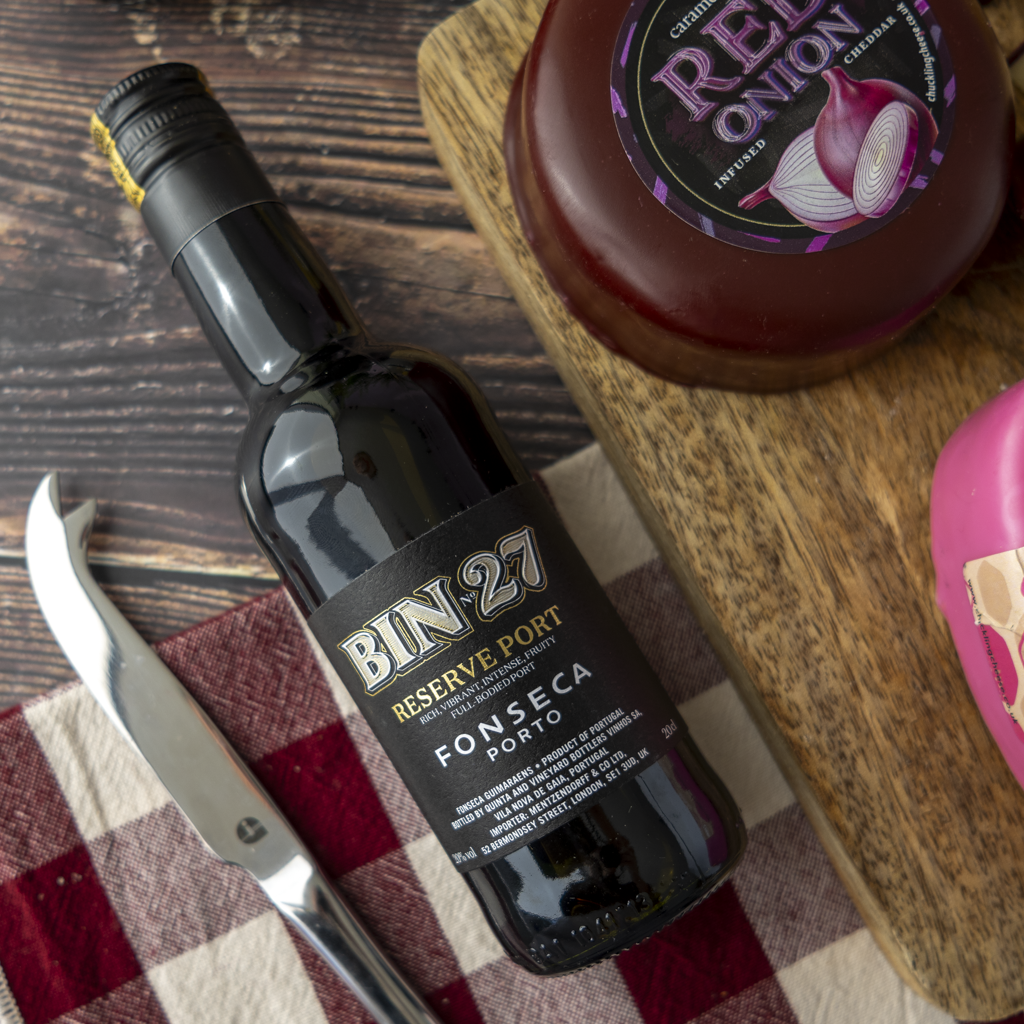 A bottle of Bin 27 Reserve Port FONSECA Porto wine styled on a rustic wooden table with a red-and-white checkered cloth, a knife, a round of red onion jam, and a pink pastry.