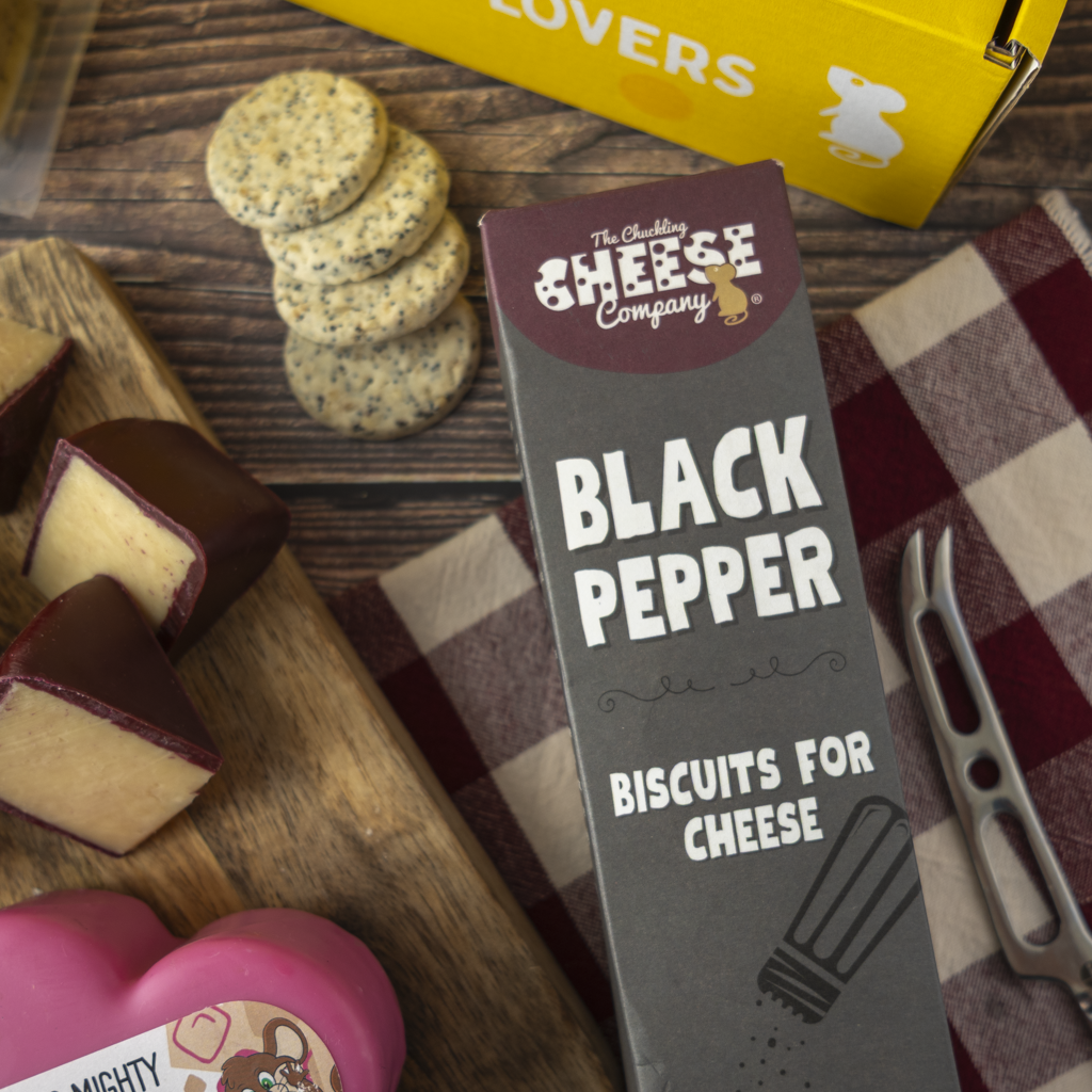 A boxed food product labelled 'The Chuckling Cheese Company Black Pepper Biscuits for Cheese' standing upright on a wooden surface, surrounded by cheese wedges, round biscuits, a yellow gift box, and a red-and-white check cloth.