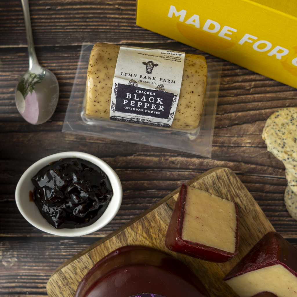 A packaged block of Lynn Bank Farm Organic Black Pepper Goudenisse cheese is displayed on a dark wood surface with a small bowl of dark pepper jam, a spoon, and sliced cheese wedges on a wooden board. A yellow box with partial white text is visible in the background.