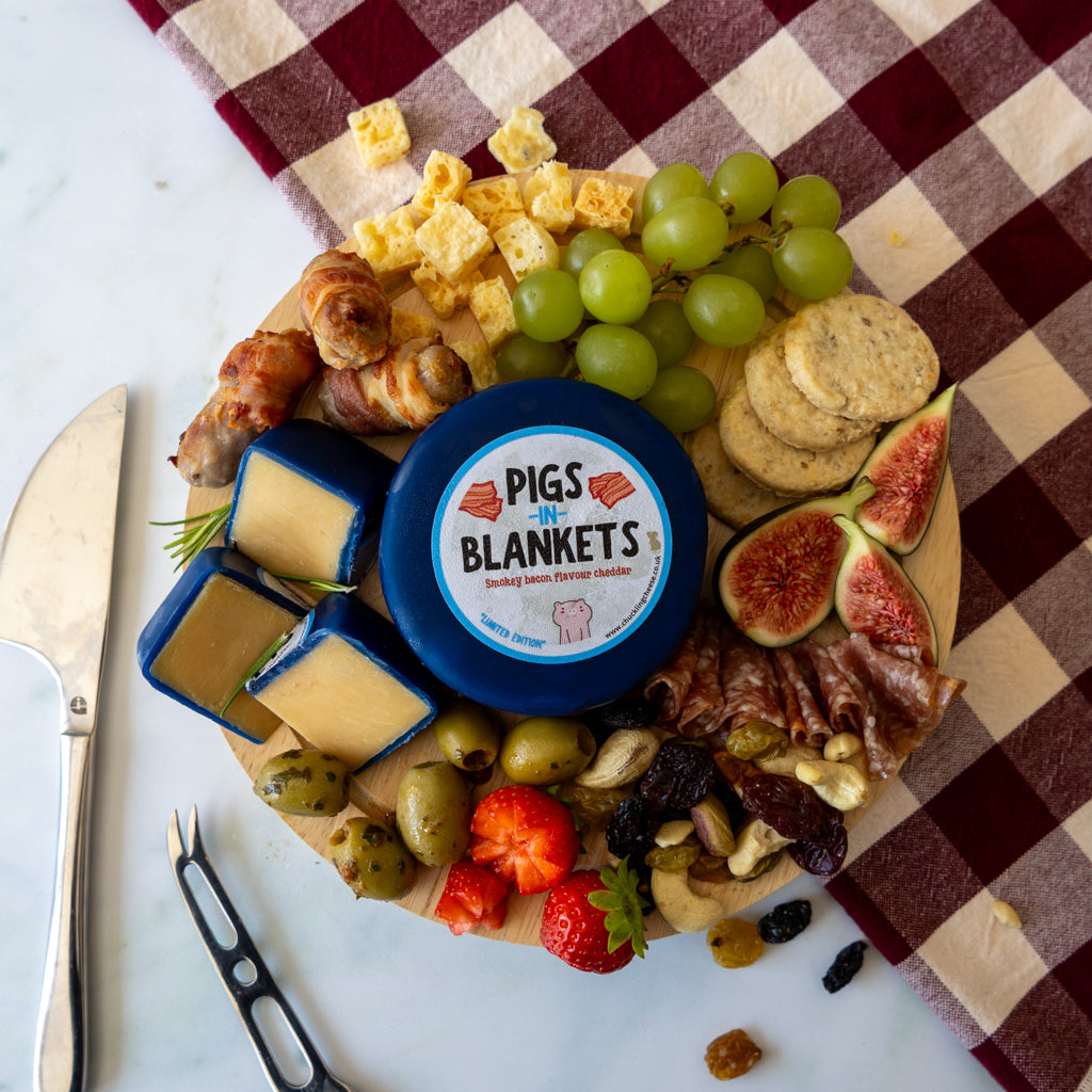 A charcuterie board styled with cheese, grapes, crackers, figs, olives, nuts, and cured meats on a round wooden board, placed on a white marble surface with a red-and-beige checkered cloth. A round blue-labeled cheese container in the center reads 'PIGS IN BLANKETS'.