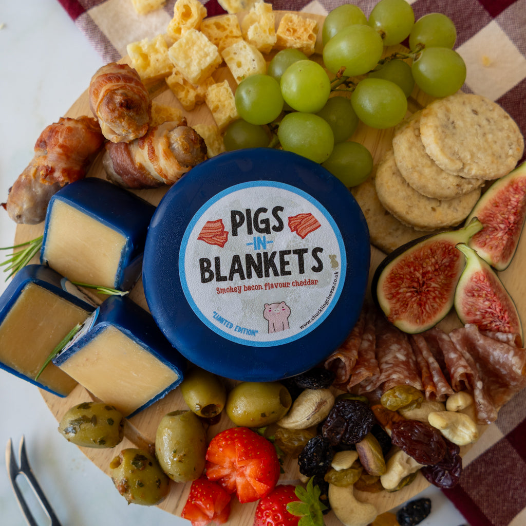 A round blue-labeled cheese wheel from Pigs & Blankets sits on a wooden charcuterie board surrounded by grapes, crackers, figs, olives, strawberries, prosciutto, salami, and cheese wedges.
