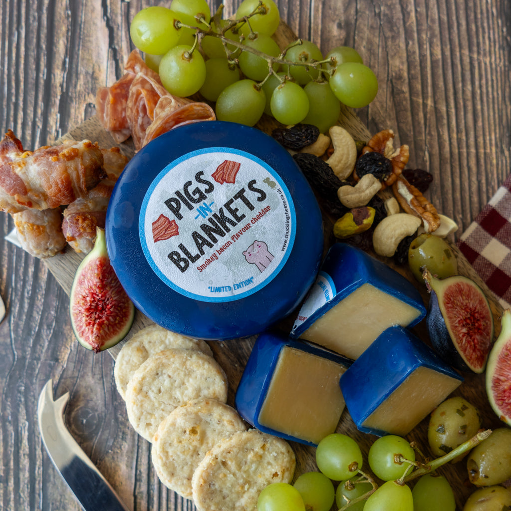 A styled food scene on a rustic wooden board featuring a round blue-labeled wheel of cheese and several wedges of the same cheese, surrounded by green grapes, figs, crackers, cured meat, nuts, and dried fruit. The cheese label reads 'PIGS ON BLANKETS'.