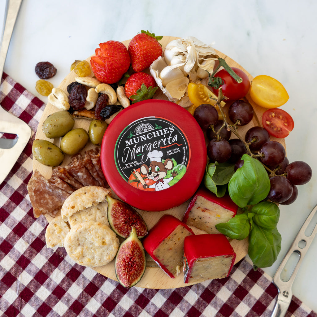 A round cheese wheel labeled MUNCHIES Margarita sits on a charcuterie board surrounded by strawberries, grapes, olives, crackers, figs, mushrooms, salami, and basil on a red-and-white checkered cloth.
