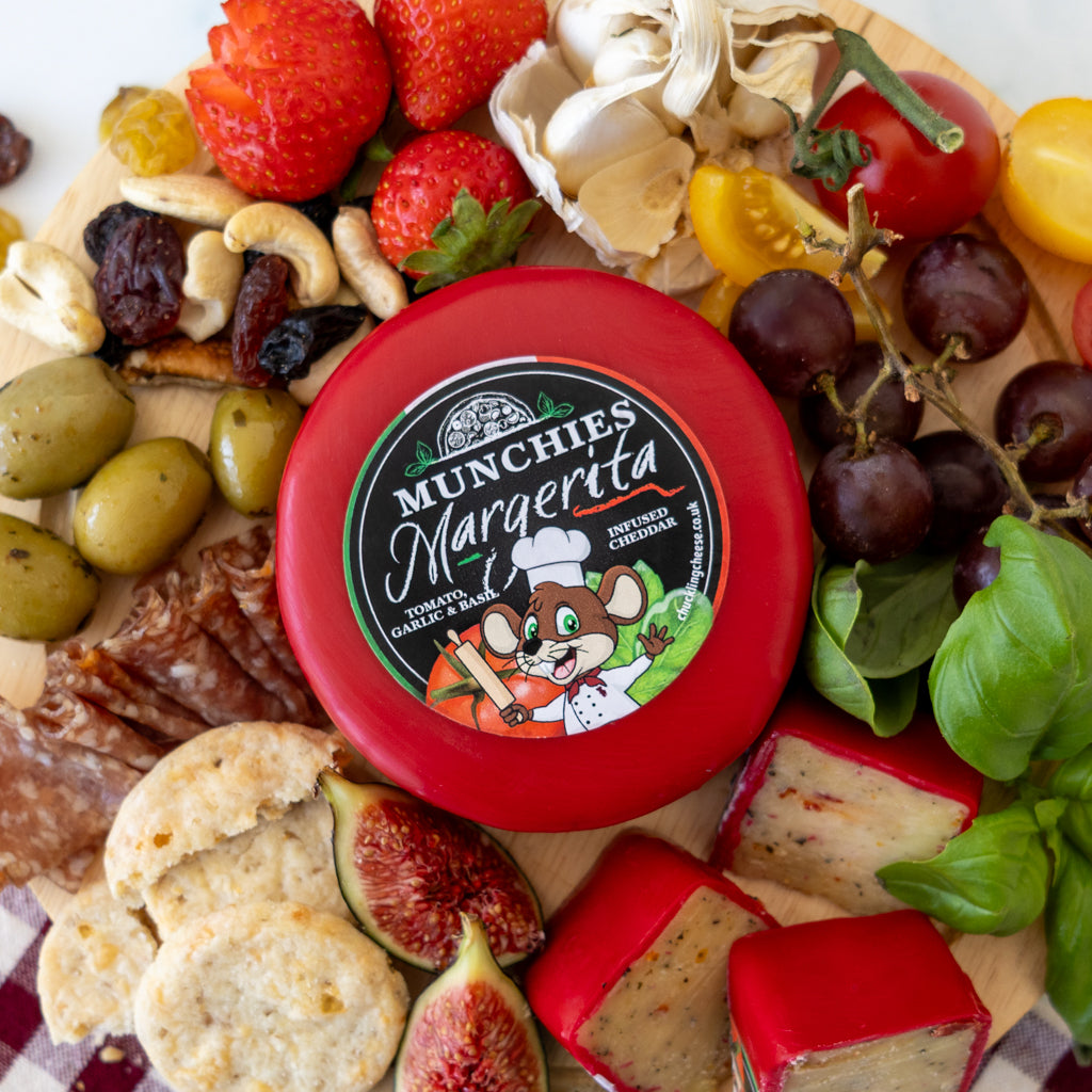 A round red-wrapped cheese wheel labeled 'MUNCHIES Margarenta' sits on a wooden charcuterie board surrounded by sliced figs, grapes, olives, crackers, basil, mushrooms, and other cheeses.