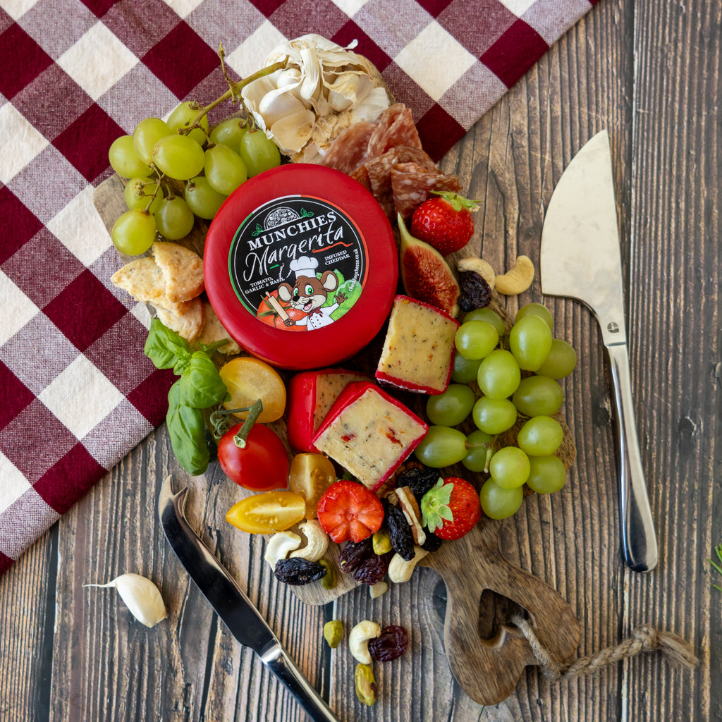 A round red-lidded jar of Munchip's Margarita cheese is arranged on a wooden board with grapes, crackers, prosciutto, basil, strawberries, nuts, and knives, set on a rustic wood surface with a red-and-white check cloth.