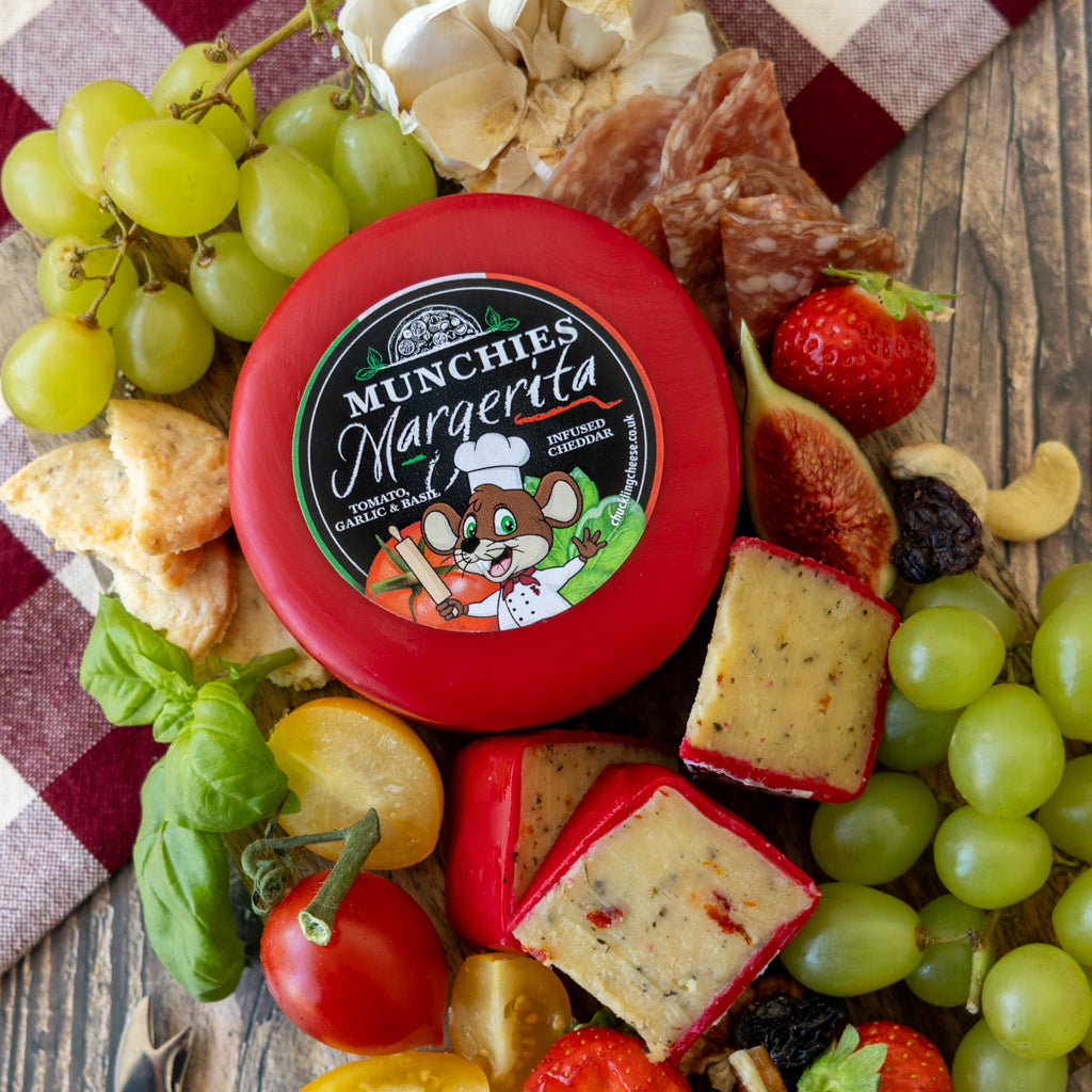 A round red container of Munchies Margarita cheese is surrounded by an appetizing charcuterie-style arrangement on a rustic wooden surface, including green grapes, basil, tomatoes, figs, crackers, salami, garlic, and a red-and-white check cloth.