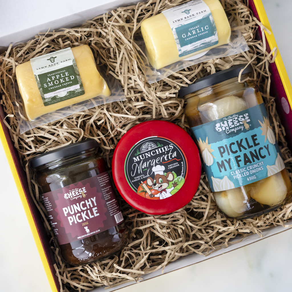 A curated cheese gift box filled with shredded paper, containing five items from Chuckling Cheese Company: two whole cheese wheels, two glass jars of pickles, and one round tin of margerita. Visible labels include Apple Smoked, Garlic, Punchy Pickle, Pickle My Fancy, and Munchies Margarita.