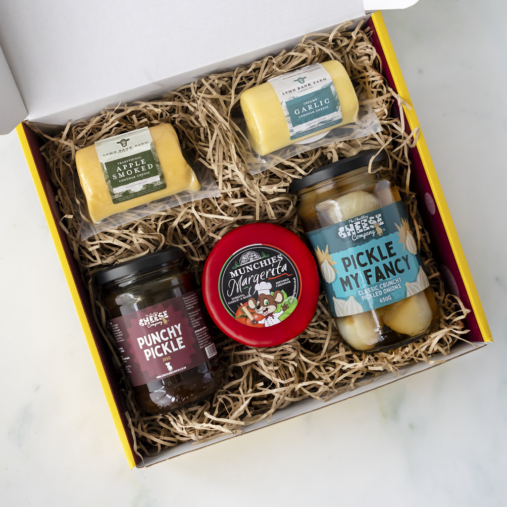 A cheese and pickle gift box from The Chuckling Cheese Company, featuring four items nestled in brown paper shred: two cheeses (Apple Smoke and Garlic), a jar of Pickle My Fancy, and a jar of Punchy Pickle, all in a yellow and white box.
