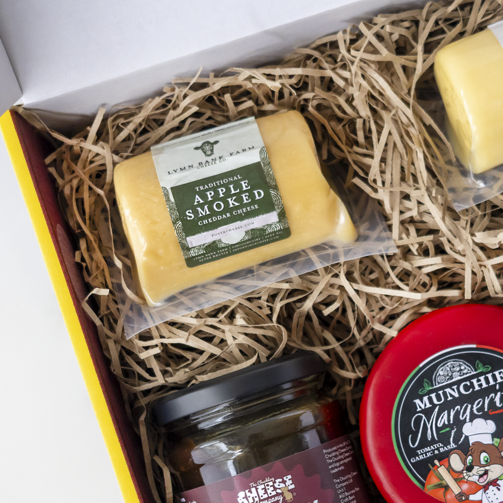 A curated cheese gift box filled with brown paper shred, featuring a package of Lynn Bank Farm Apple Smoked Cheddar Cheese, a jar of cheese spread, and a red-labeled Munchie Margherita cheese wheel.