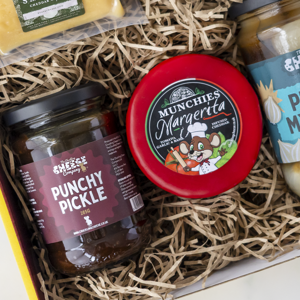 A curated gourmet gift box filled with shredded paper, featuring a jar of The Chuckling Cheese Company Punchy Pickle, a round wheel of Munchies Margarita cheese, and other gourmet items partially visible.