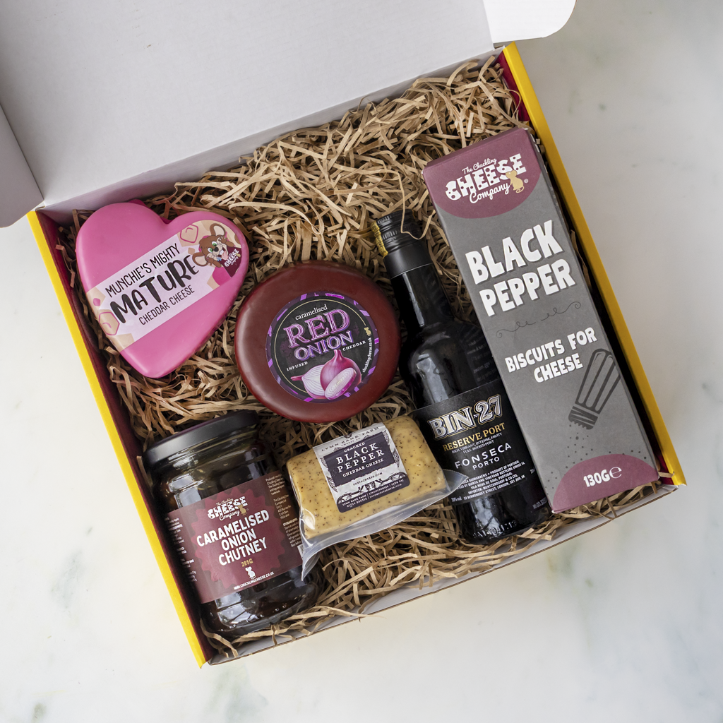 A curated cheese gift box filled with shredded brown paper, containing a pink heart-shaped Mature cheese truckle, a round Red Onion cheddar cheese wheel, a jar of caramelised onion chutney, a box of crackers, and a bottle of BIN27, all arranged in a white box with a yellow border on a light marble surface.