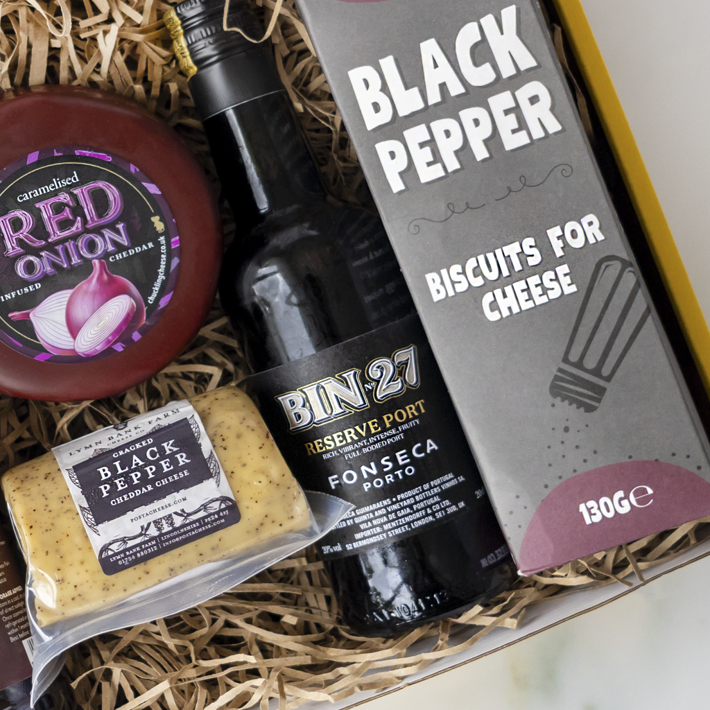 A curated cheese and snack gift box filled with shredded paper. Visible items include a Bin-27 Reserve Port Fonseca Porto bottle, a package of Black Pepper Biscuits for Cheese, a round Red Onion cheddar cheese wheel, and a packaged Black Pepper Cheddar Cheese barrel.
