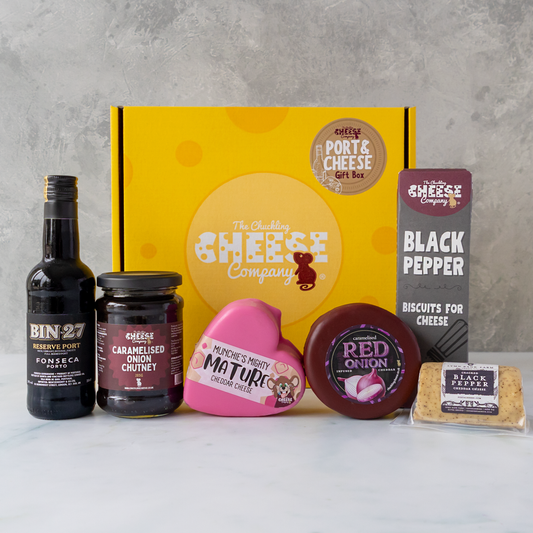 A curated cheese and food gift box set from The Chuckling Cheese Company, featuring a yellow gift box, a bottle of Bin27, caramelised onion chutney, a heart-shaped Mature cheese truckle, a round Red Onion cheese truckle, and a pack of Biscuits for Cheese Black Pepper biscuits, arranged on a white marble surface against a grey concrete wall.