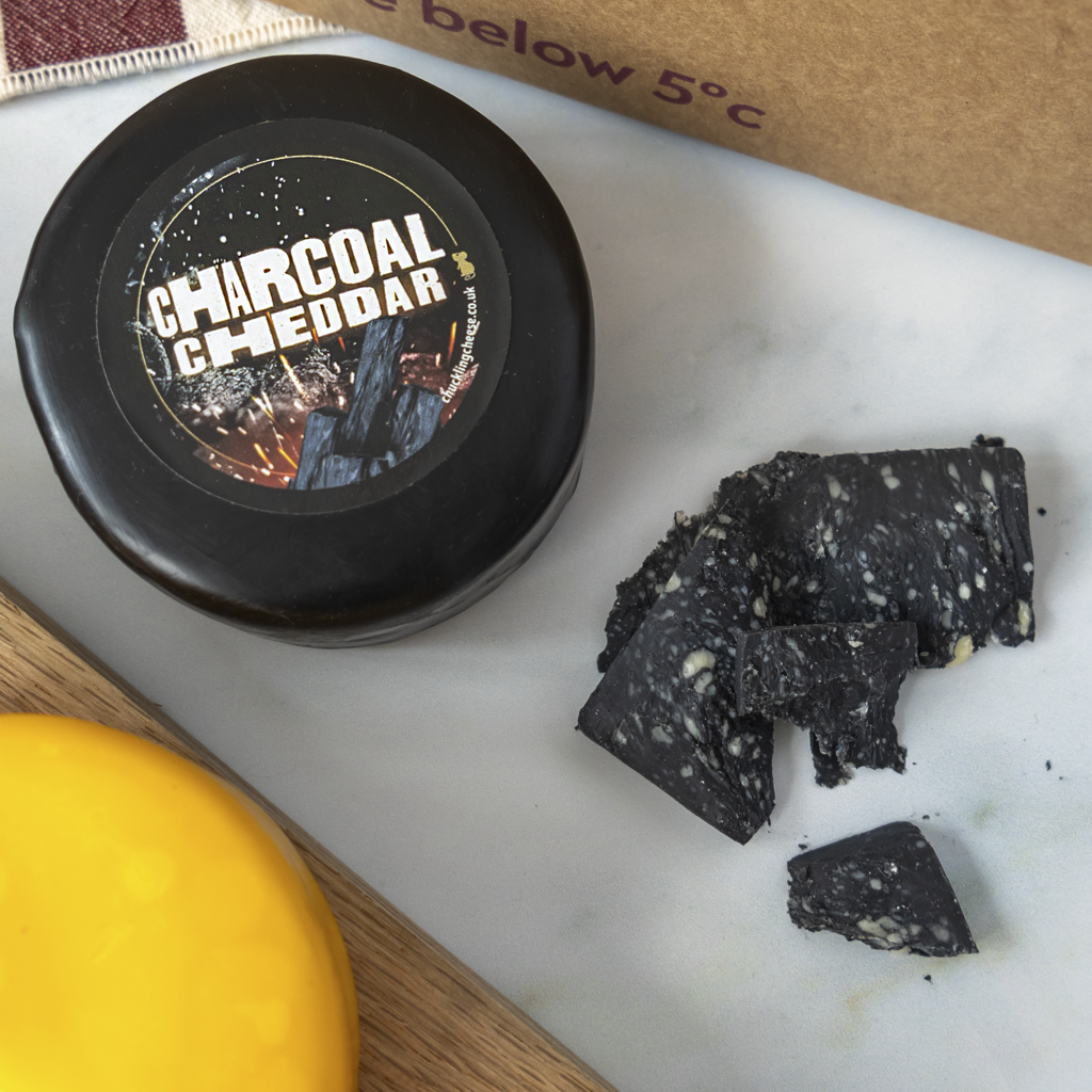 A round black cheese truckle labeled 'CHARCOAL CHEDDAR' sits on a white marble surface beside broken pieces of dark charcoal-colored cheddar cheese and a bright yellow cheese wedge, with a brown paper bag partly visible in the background.