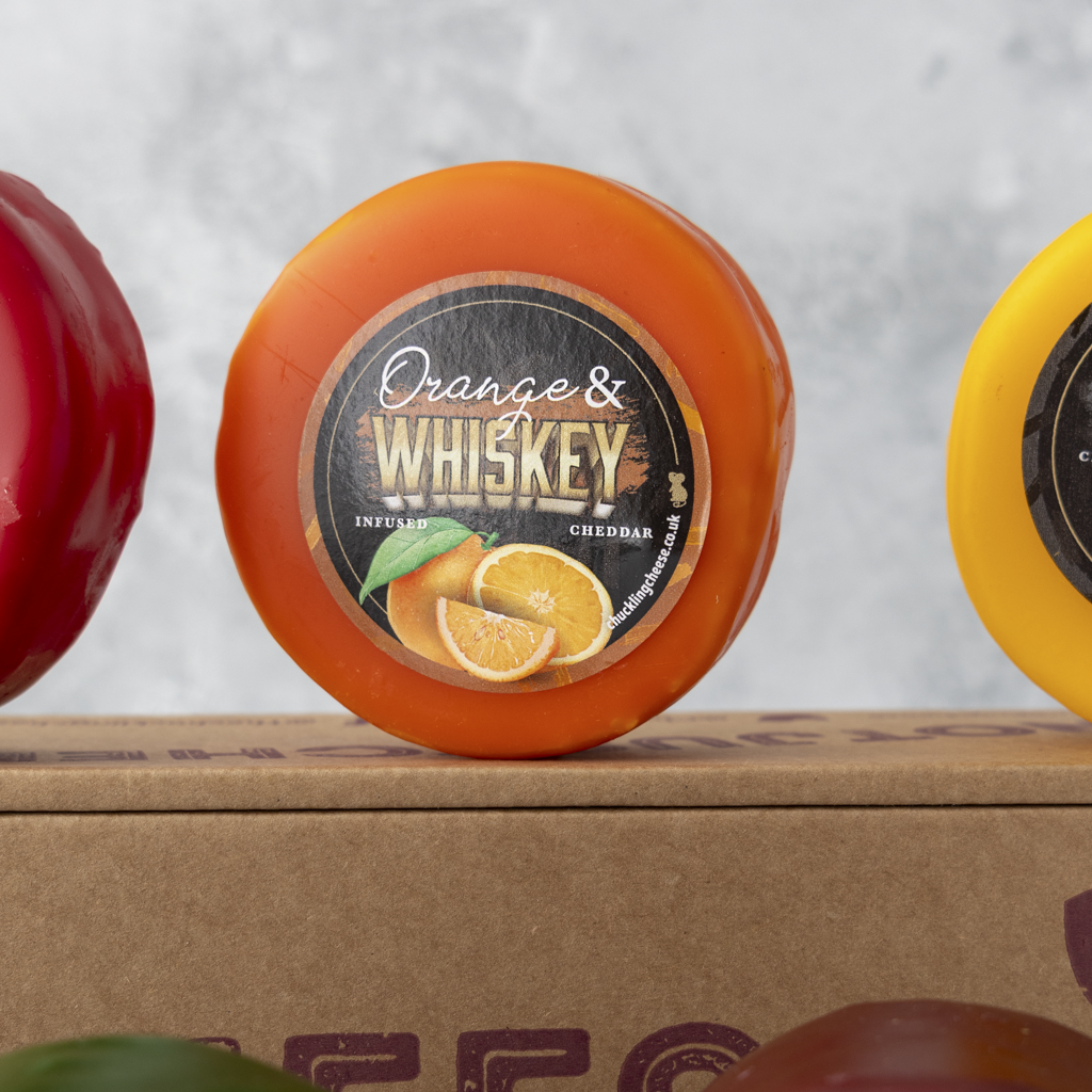 An orange cheddar cheese wheel labeled 'Orange & Whiskey' sits on a cardboard box, with red and yellow cheese wheels partially visible in the background.