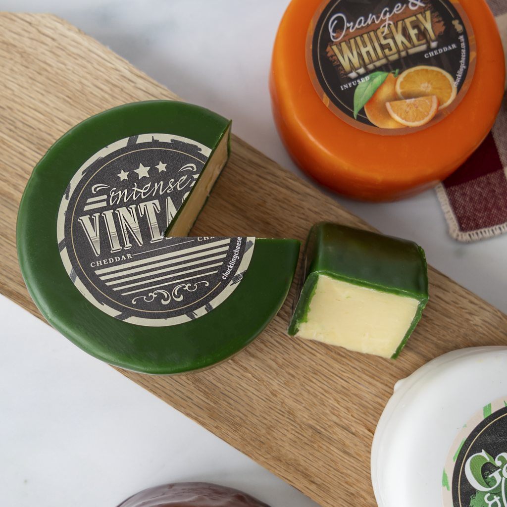 A styled product shot of artisanal cheeses on a wooden board. The main focus is a dark green-wrapped cheese wheel labeled 'Intense Vinta Cheddar' with a wedge cut out and a matching cut piece beside it.