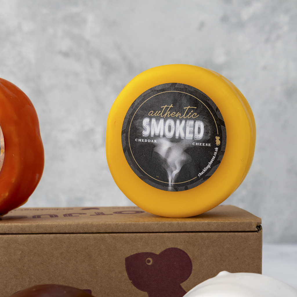 A round wheel of smoked cheddar cheese with a bright yellow rind and a black circular label reading 'authentic SMOKED cheddar cheese' sits on top of a brown cardboard box. Part of another orange cheese wheel is visible on the left, and a white cheese wheel is partially visible at the bottom right, against a light gray textured background.