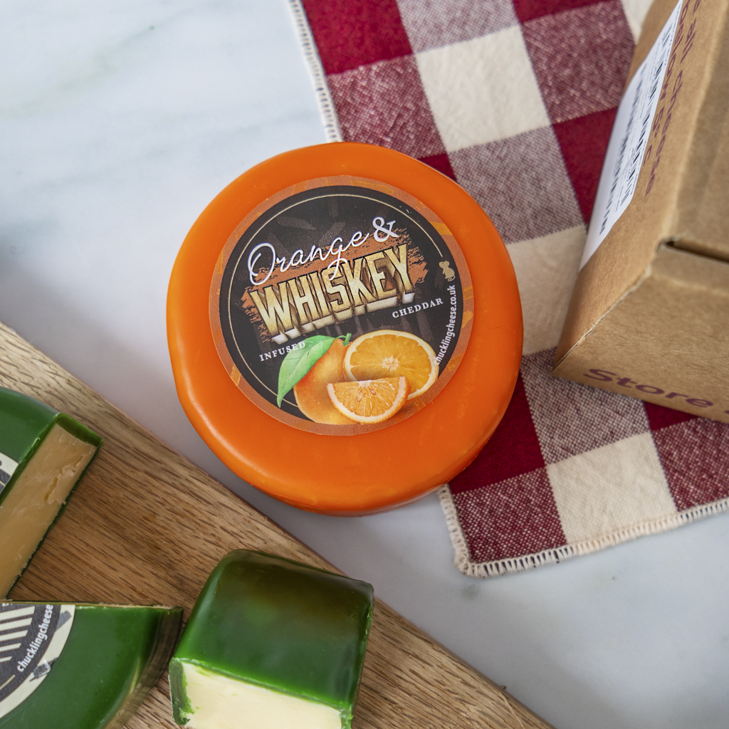 A round orange cheese wheel labeled 'Orange & Whiskey' is shown on a marble surface with a red-and-white check cloth, a kraft shipping box, and cheese wedges.