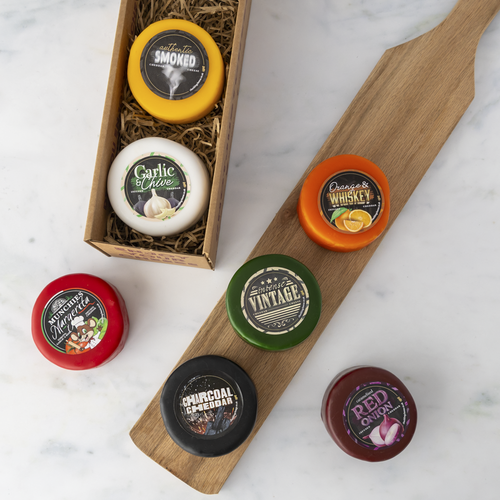 A flat-lay of five round cheese wheels with vintage-style labels arranged on a wooden serving board and a small wooden gift box filled with shredded paper, on a light marble surface.