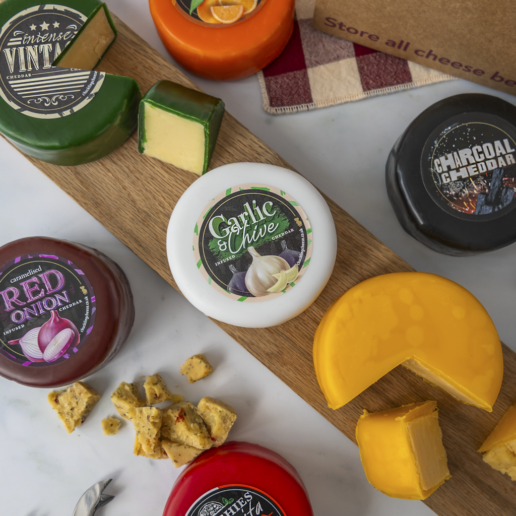 A styled flat-lay of assorted artisan cheeses on a wooden board and white surface, including a green wheel labeled Vintage, a white round labeled Garlic Chive, a black wheel labeled Charcoal Cheddar, a red jar labeled Red Onion, and a bright orange cheese wedge with croutons.