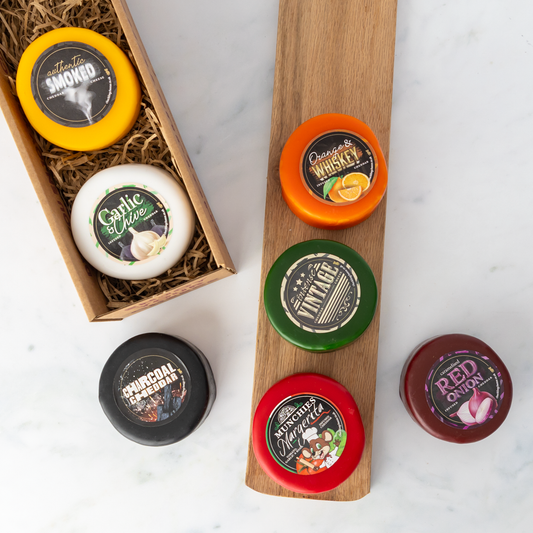 Six small round cheese wheels with colorful labels are arranged on a wooden display board and in a wooden gift box on a white marble surface. The labels include flavours such as Orange Whiskey, Garlic Chive, Charcoal Cheddar, and Red Onion.