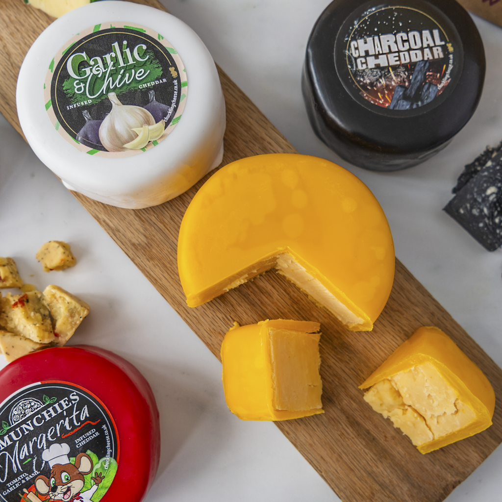 A styled cheese assortment on a wooden board, featuring a sliced round yellow cheddar cheese, a white round cheese labeled Garlic & Chive, a red round cheese labeled Munchies Margarita, and a black cheese truckle labeled Charcoal Cheese.