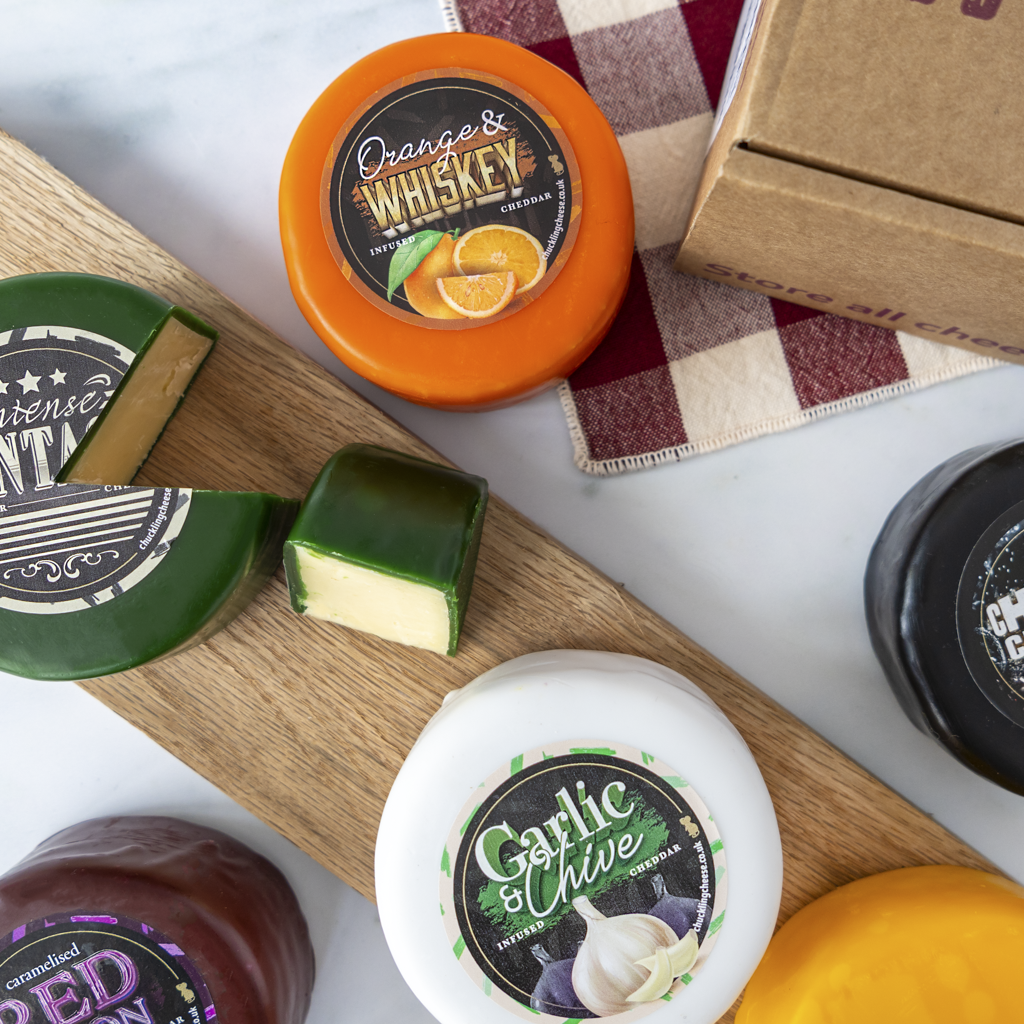 An overhead styled flat-lay of several artisanal cheese wheels and a sliced wedge on a wooden board, with one orange cheese wheel labeled Orange & Whiskey, one white-lidded wheel labeled Garlic & Chive, and a kraft box partially visible on a white marble surface.