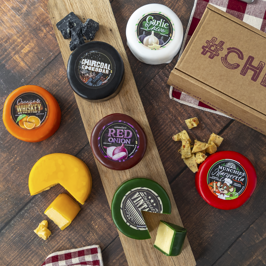 A curated cheese and snack board arranged on a dark wood surface, featuring several round cheese wheels with cut wedges, a wooden serving board, crackers, and a brown gift box with '#CHEESE' printed on it.