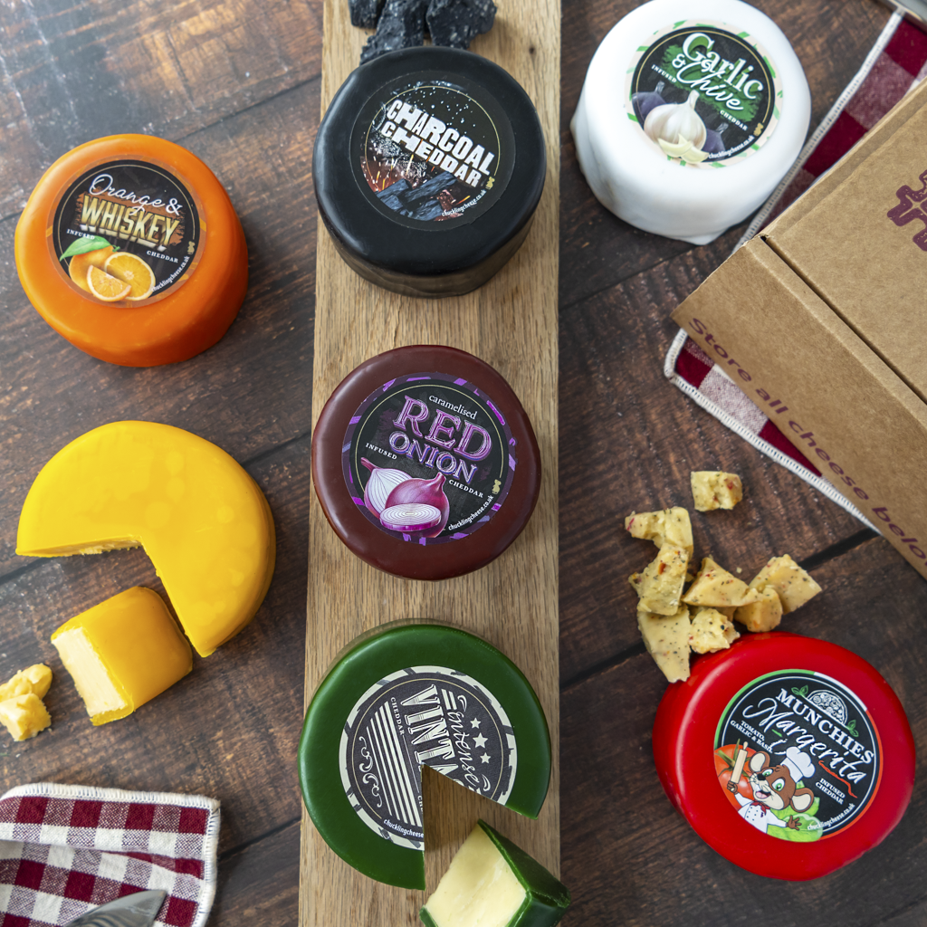 A styled flat-lay of artisanal cheese truckles on a dark wood background, including several round cheese truckles with colourful lids, a wedge of yellow cheese with a slice cut out, crackers, and a cardboard box. Visible labels include Orange & Whisky, Charcoal Cheddar, Garlic & Chive, and Munchies Margerita.