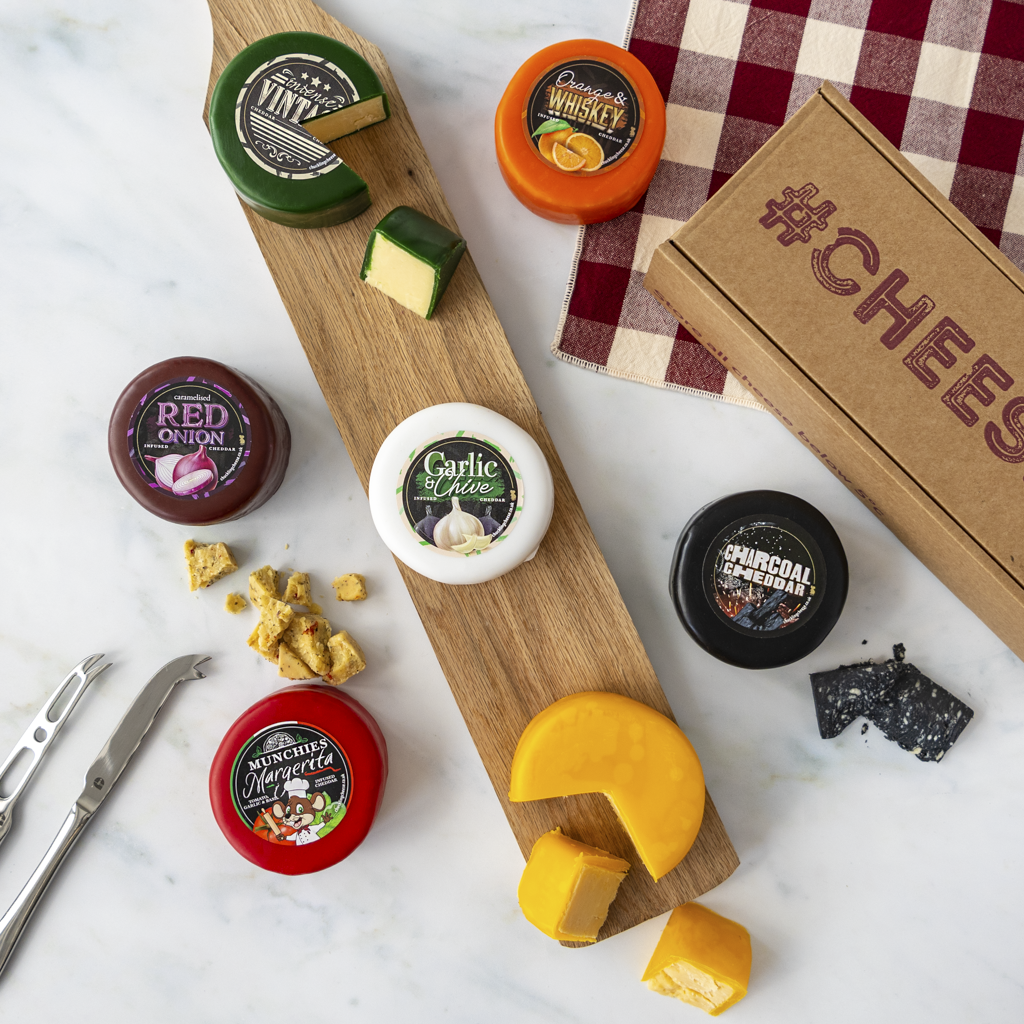 A curated cheese truckle assortment arranged on a marble surface with a wooden serving board, including multiple round cheeses with printed labels, a wedge of yellow cheese, crackers, cheese knives, and a cardboard box marked “#CHEESE.”.