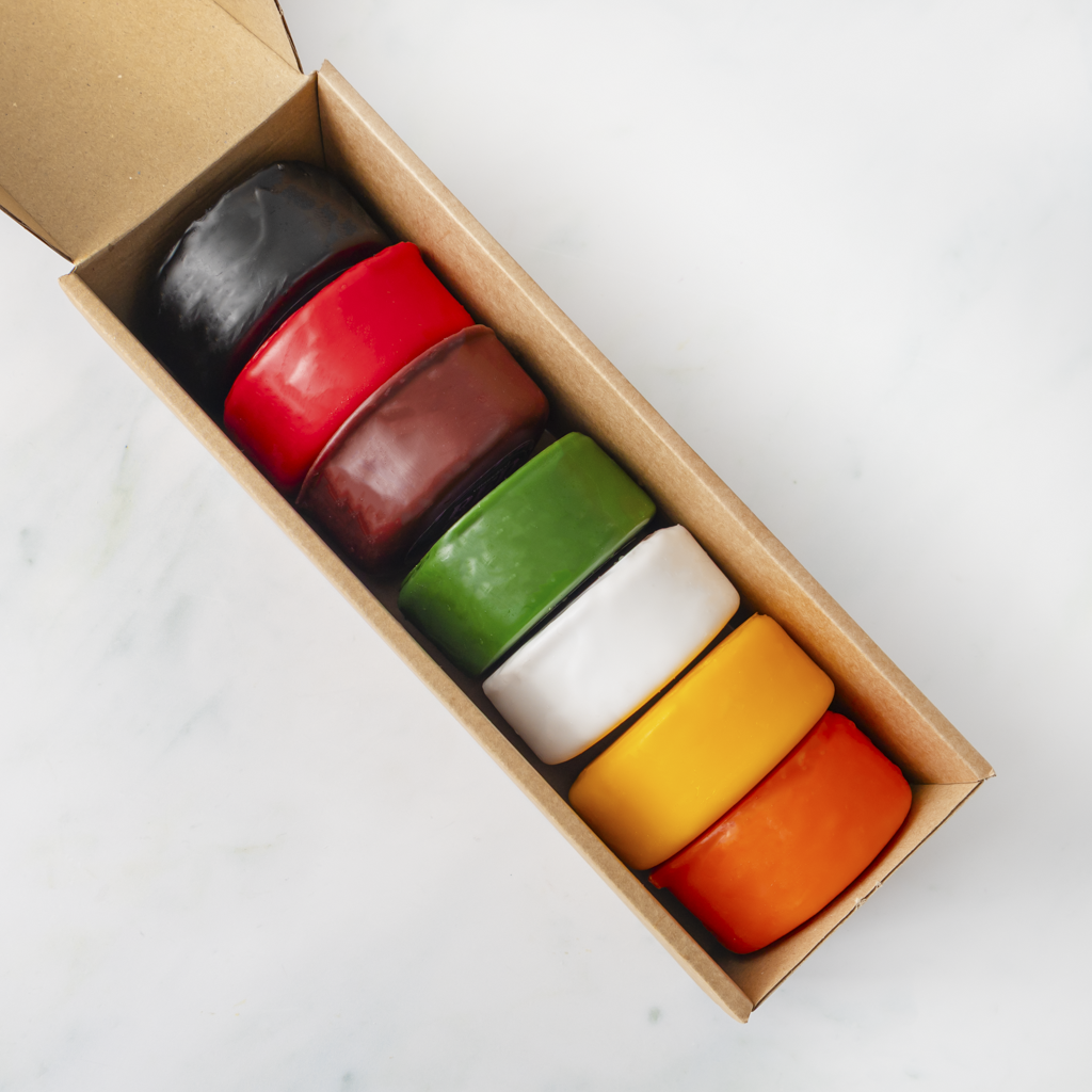Seven colourful circular cheese truckles stacked inside an open brown cardboard box on a light marble-like surface. The colours visible are black, red, dark brown, green, white, yellow, and orange.