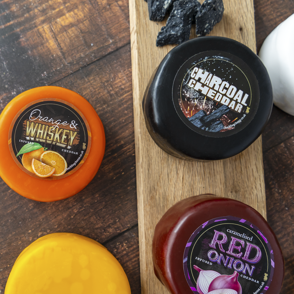 Four gourmet cheddar cheese wheels arranged on a wooden board. The labels read Orange & Whiskey, Charcoal & Cheddar, and Red Onion.