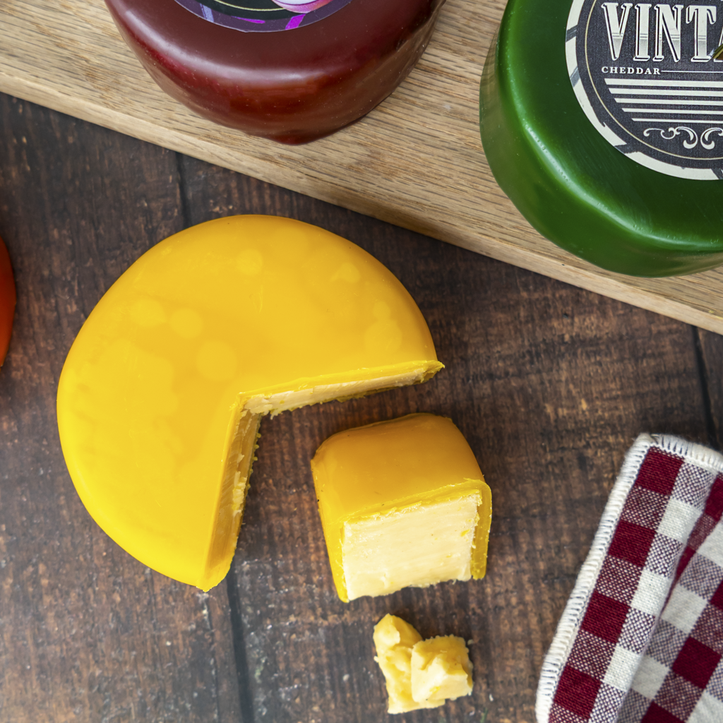 A wedge of yellow cheddar cheese with a slice cut out, showing the creamy pale interior, set on a dark wooden surface with a red-and-white check cloth and partial views of other cheese wheels in the background.