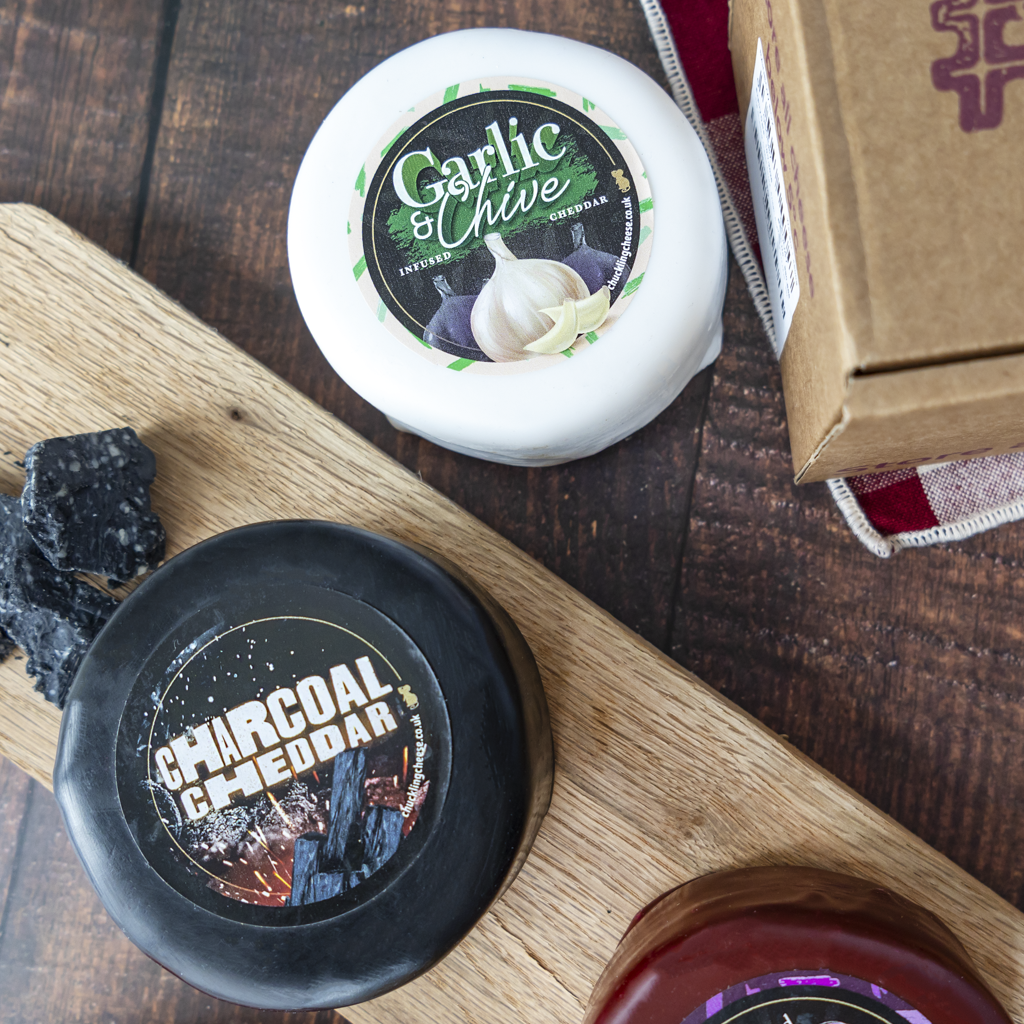 A styled flat-lay of gourmet food products on a dark wood background, featuring a round white cheese labeled Garlic & Chive, a round black cheese labeled Charcoal Cheddar on a wooden board, a bagel, and a brown cardboard box.