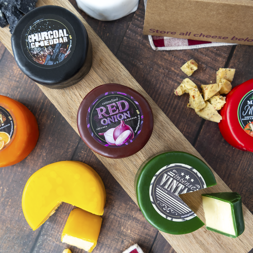 A styled cheese assortment on a wooden board and table, including round cheeses with labels for Charcoal Cheddar, Red Onion, and Intense Vintage, plus sliced cheeses and cheese pieces.