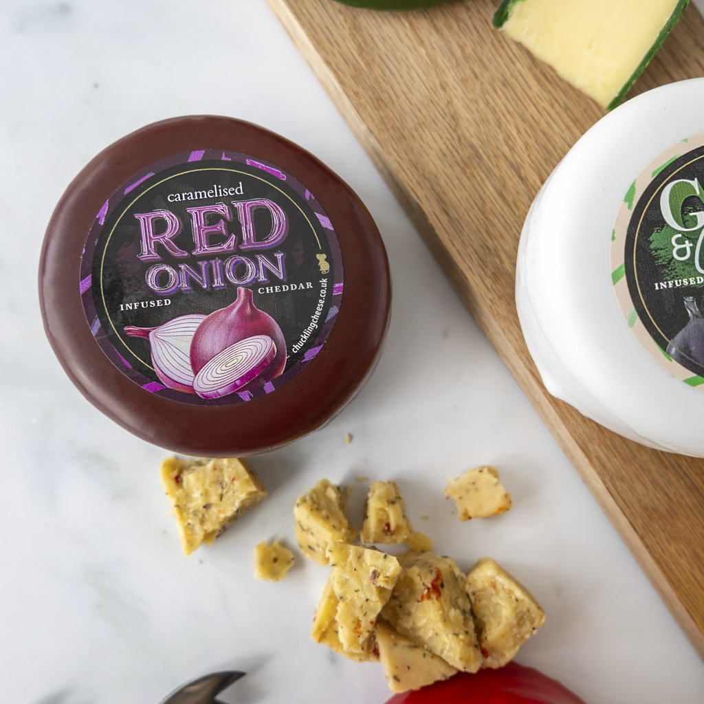 A round wheel of caramelised red onion cheddar cheese with a dark reddish-brown rind and a black label reading 'RED ONION' is shown on a white marble surface. Sliced cheese pieces are scattered in front, with another cheese wheel and a wooden cutting board partially visible in the background.