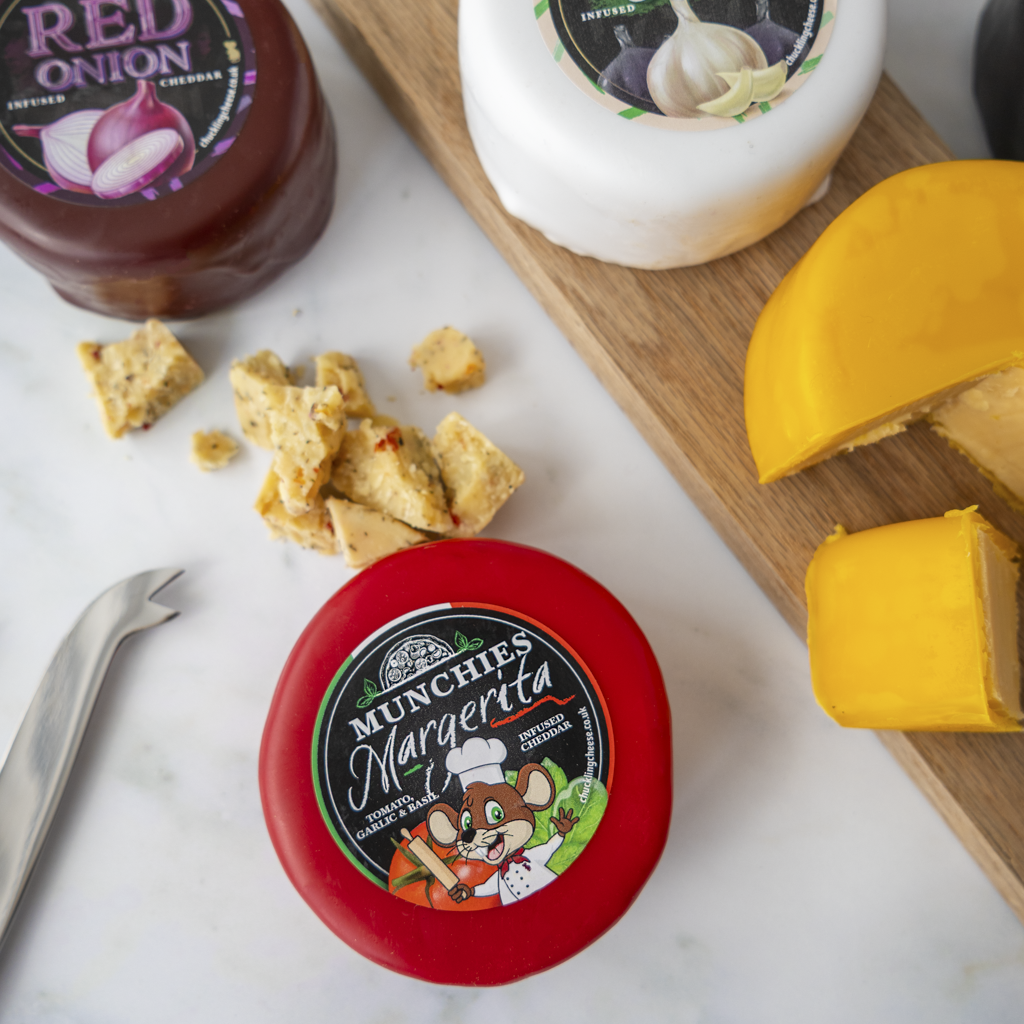 A round red container of Munchies Margarita cheese dip is shown on a white marble surface with crackers, a wedge of yellow cheese, a white wheel of cheese, and a bottle of red onion dip.