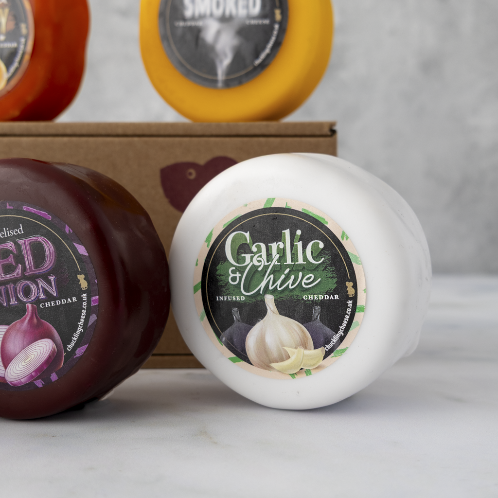 Three round cheese wheels with colourful labels are arranged on a light marble surface in front of a brown cardboard box. The front-right wheel is white with a black, green, and beige 'Garlic & Chive' label; the left wheel is dark red with a purple onion label; the top background wheels are orange and yellow with smoked labels.