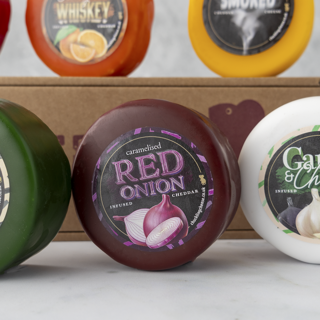 A round red onion cheddar cheese wheel in the foreground, with other cheese wheels and a cardboard box blurred in the background. The central label reads 'caramelised RED ONION INFUSED CHEDDAR'.