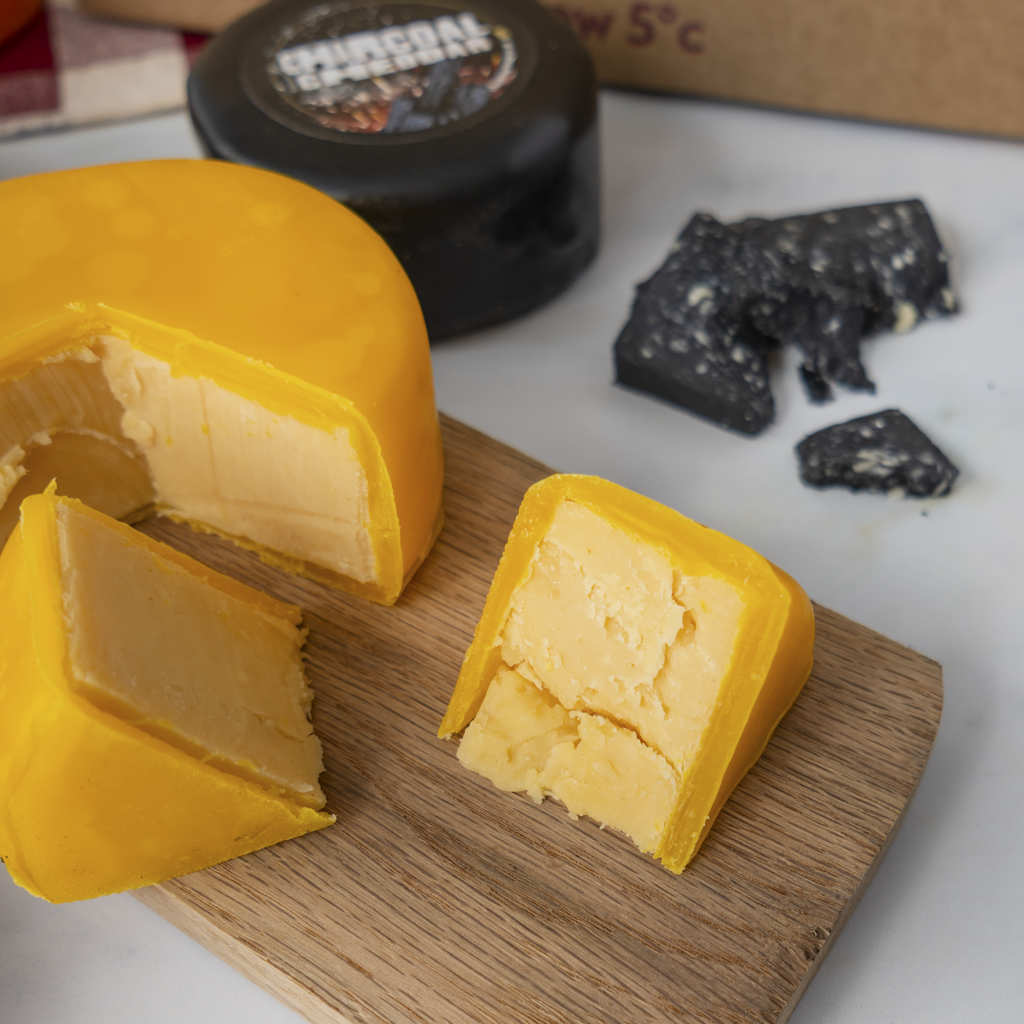 A close-up of a yellow cheese wheel and wedges on a wooden board, with a dark round container and broken dark cheese pieces in the background on a white surface.
