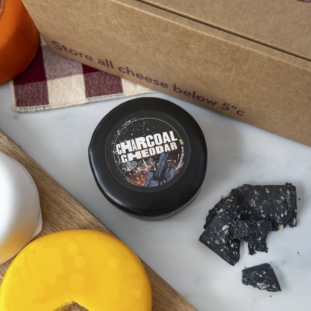 A round black container of Charcoal Cheddar cheese sits on a white marble surface with cheese slices and a brown cardboard box nearby. The label reads 'CHARCOAL CHEDDAR'.