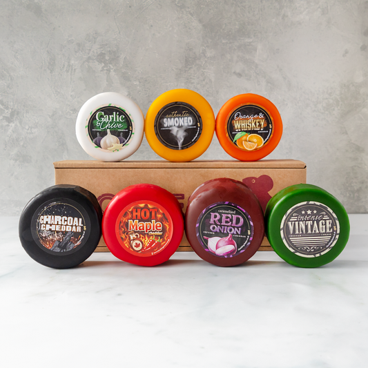 Seven round wax melts in assorted cheese truckle flavours are arranged on and around a brown cardboard box against a light grey textured wall and white surface. The cheese include Garlic & Chive, Smoked, Whiskey, Charcoal, Hot Maple, Red Onion, and Vintage.